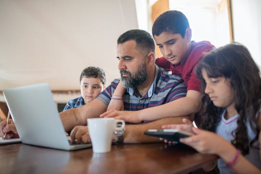 A father manages work tasks at home while bonding with his children around a table.