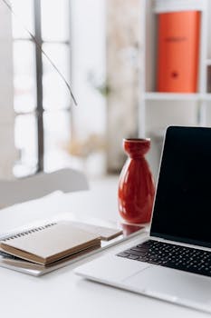 A minimalist home office setup featuring a laptop, notebook, and a red vase on a white desk.