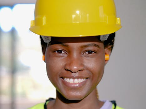 Close-up of a smiling female engineer wearing a yellow hard hat and ear protection indoors.